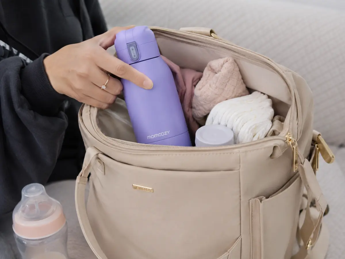 The Momcozy portable bottle warmer packed in a diaper bag, showing its size and the locking lid